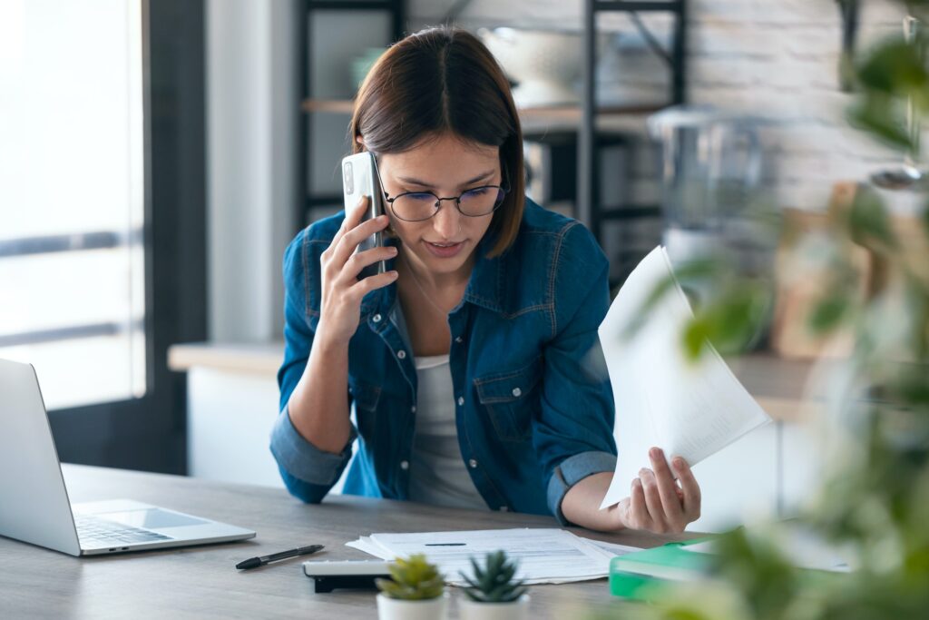 young woman talking with her mobile phone while working with computer in the kitchen